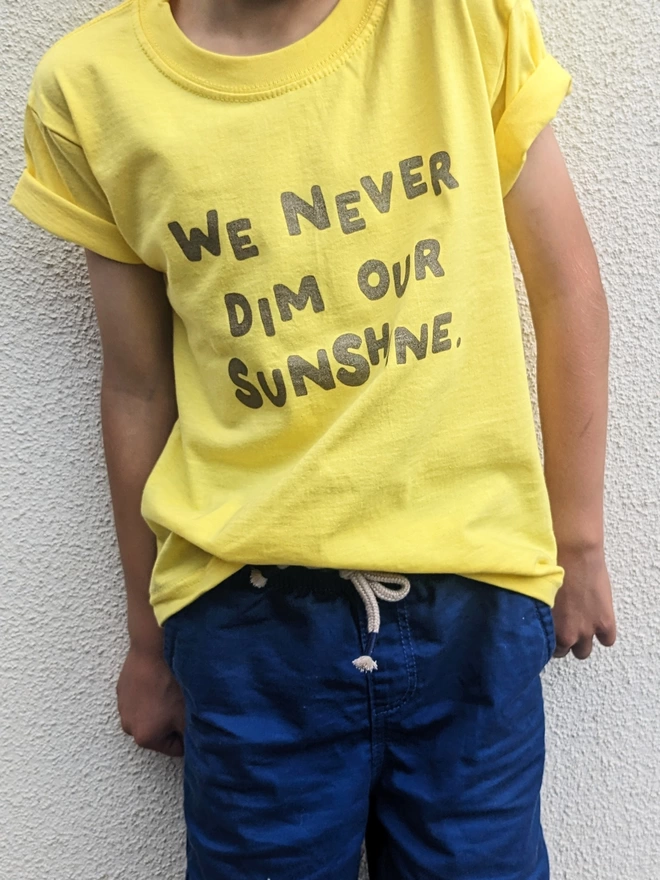A child by a white wall wearing a yellow t-shirt with We Never Dim Our Sunshine t-shirt