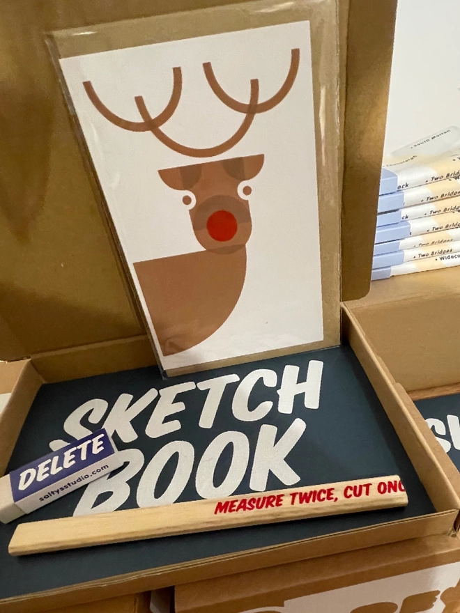 An open cardboard box with a sketchbook, carpenters pencil a Delete rubber with graphic screenprinted words upon, plus a reindeer card stood looking back at you!