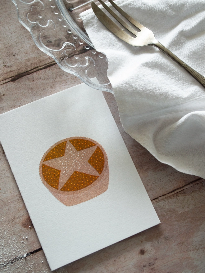 A small festive Christmas crd printed with an illustrated Mince pie lies on a wooden Christmas table beside and empty plate and desert fork.