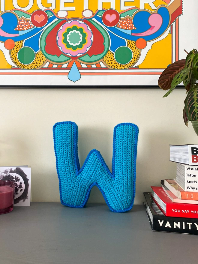 Crochet cushion shaped like the letter W standing on a shelf, in Light Blue and Blue