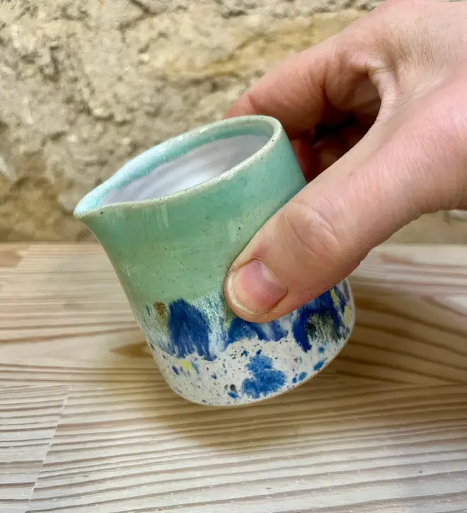 Shoreline Tiny Jug, a colourful tiny jug placed on a wooden surface. It sits in front of a brick wall backdrop. In the image its being poured. 