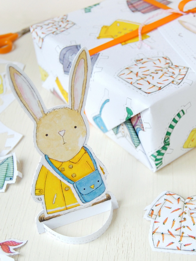 A gift wrapped in white wrapping paper with a hand illustrated outfit design and rabbit paper doll gift tags is beside a cut out rabbit paper doll on a white wooden table.