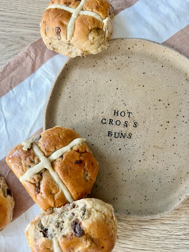 Hot cross buns speckled stoneware plate, the plate has a wavy rim and is stamped with the wording in black.