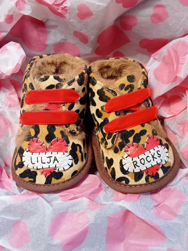 A pair of leopard print baby booties, lined with fur and with red velvet ribbon laces. A red heart with a white scroll adorns each booty. One says LILJA, one ROCKS in black stitches