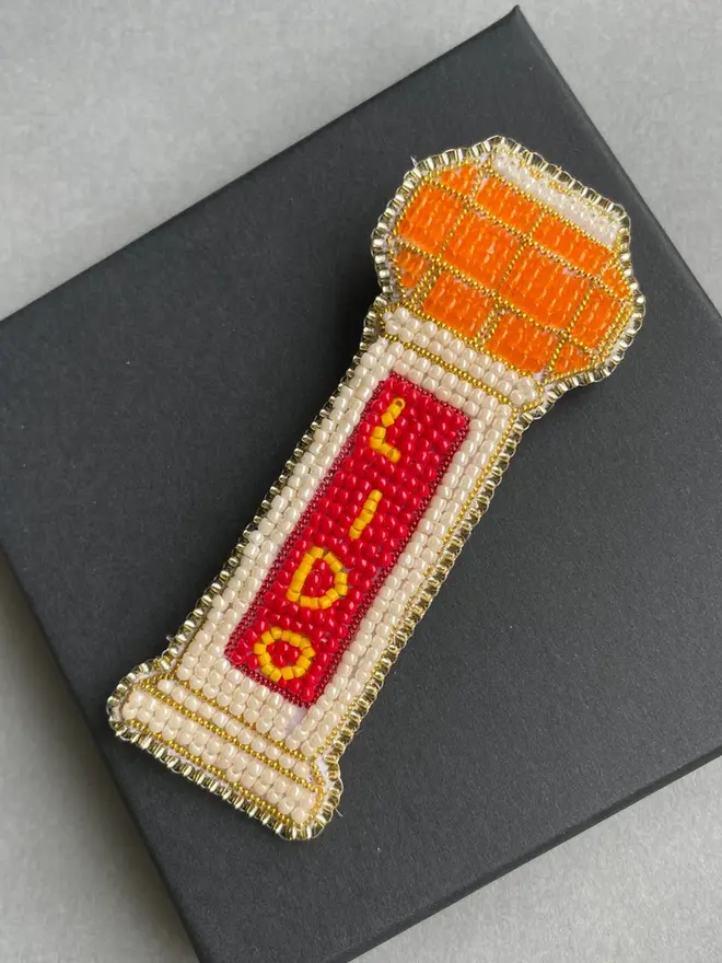 Beaded lido brooch with orange, red, and beige beads placed on top of a black box on a grey table.