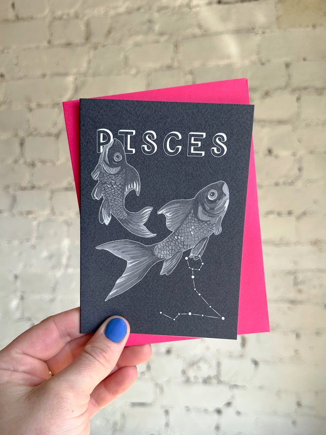 a greetings card featuring a greyscale illustration of a zodiac style couple of fish with the text “PISCES” with a neon pink envelope
