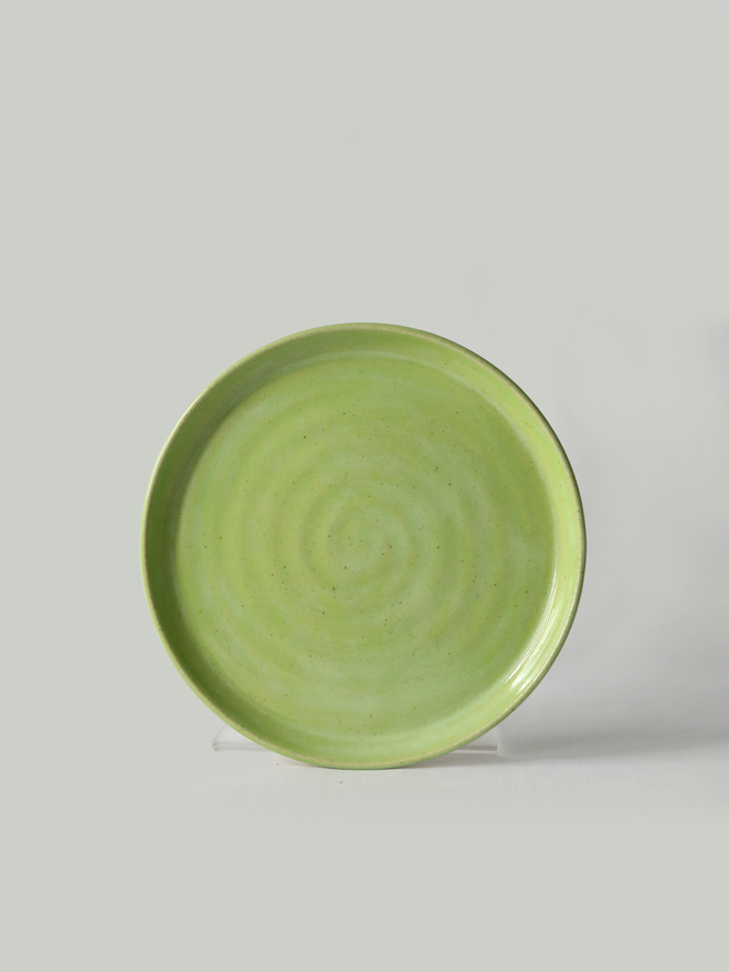 A handmade ceramic cake plate in spring green glaze, photographed from the front against a plain light grey background, showing a natural concentric swirl texture and a shallow raised rim