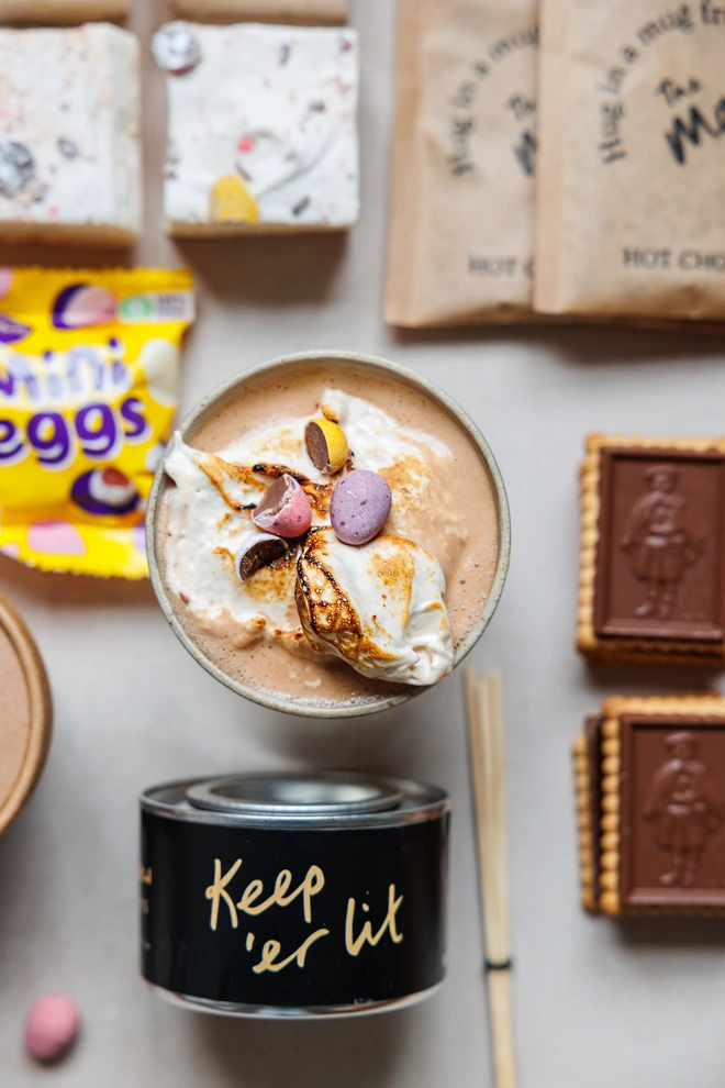 Mini Eggs Wonder Fluff close up of crushed mini eggs on top of mini eggs wonder fluff topped hot chocolate with contents of Easter luxury smores and hot chocolate kit surrounding