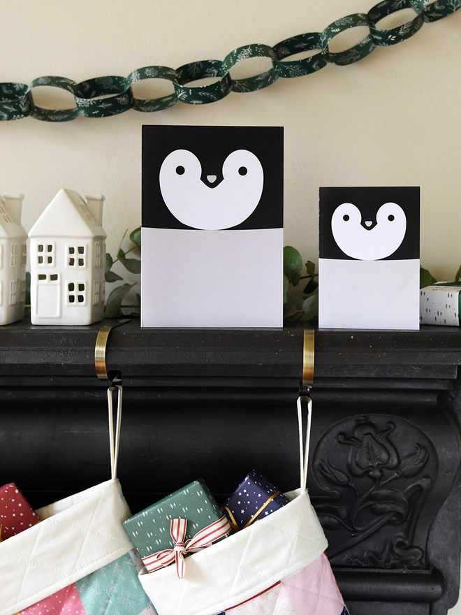 Two penguin greetings cards stand on a black mantlepiece where two patchwork stockings are hanging from.
