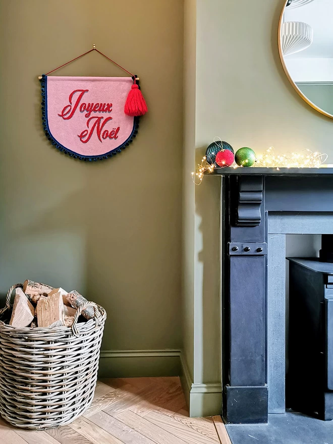 A Joyeux Noel Christmas banner hanging in a green living room next to a fireplace. A basket of logs sits on the floor beneath the wall hanging and some baubles and faity lights adorn the mantlepiece creating a warm and cosy glow.