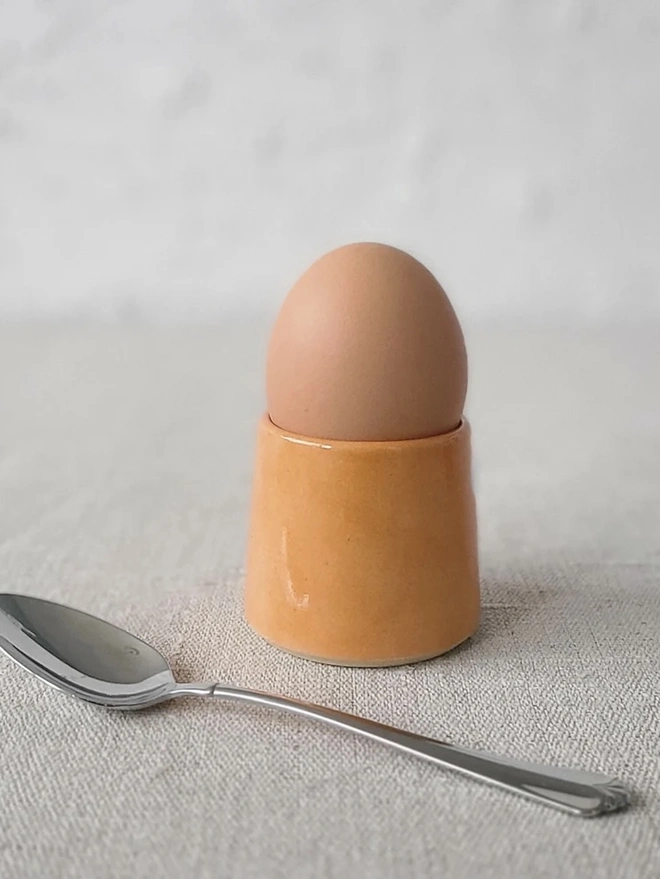 Classic Handmade Egg Cup, a ceramic egg cup sitting on a carpet surface against a grey backdrop. It is accompanied by a spoon. 