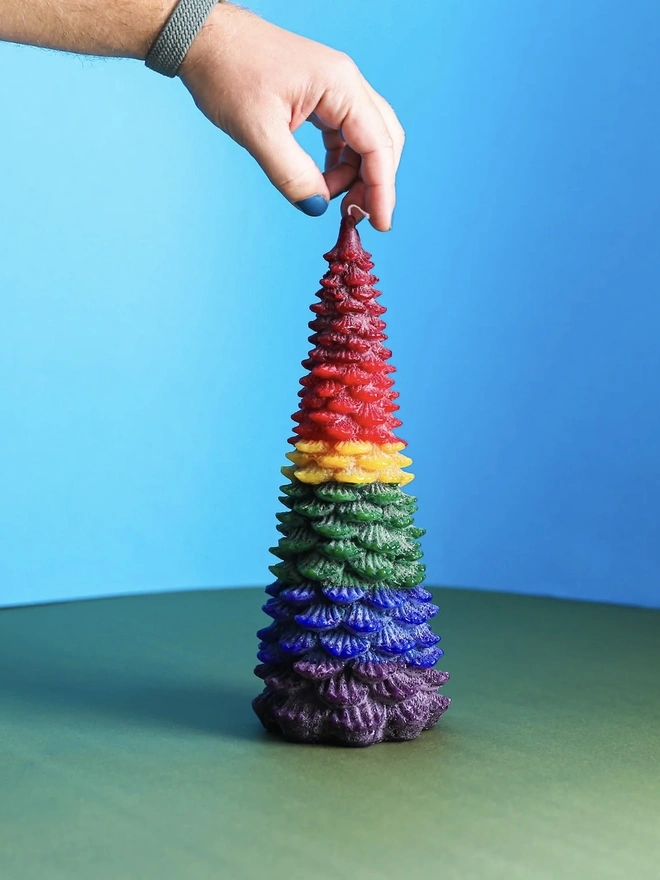 Large Christmas Tree Candle. a hand holds a large christmas tree shaped candle by its wick, showing off its rainbow ombré design in red, yellow, green, blue and purple layers. the candle stands upright against a bright blue background, with a green surface below, highlighting the textured, layered pine branch design running the full length of the tree. the multicoloured wax gives the candle a bold, playful finish.