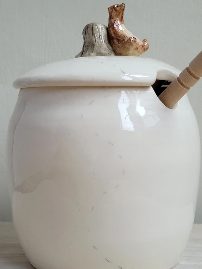 side view of a handmade ceramic white honey pot with a tiny brown bear and tree stump on the lid and a wooden honey drizzler 