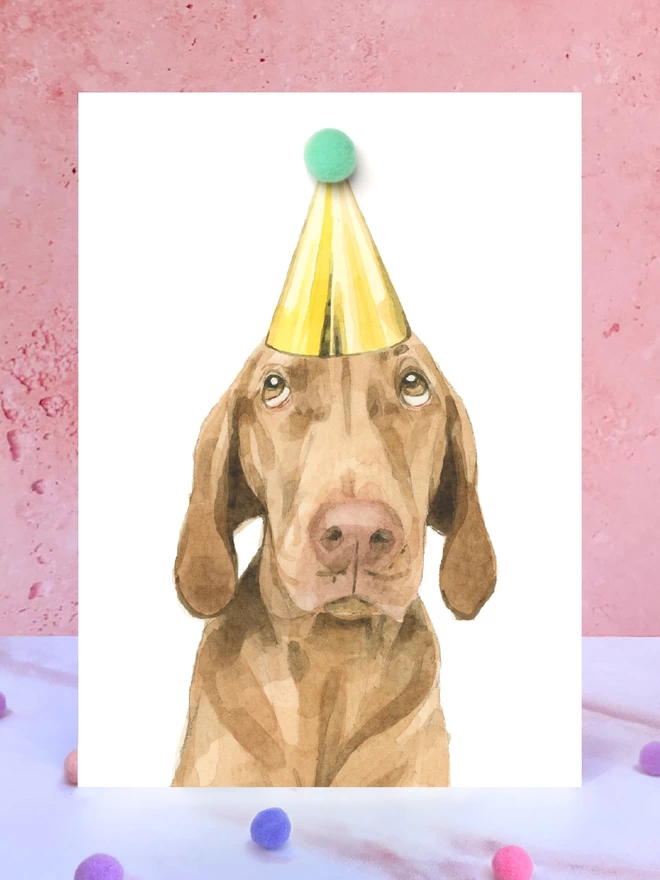 A greeting card featuring a hand painted design of a vizsla, stood upright on a marble surface surrounded by pompoms. 