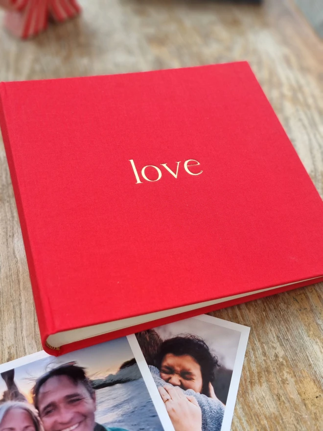 A hand-bound red photo album printed with the word 'love' in gold lettering rests on a fur rug and a wooden floor. It is surrounded by family photographs.
