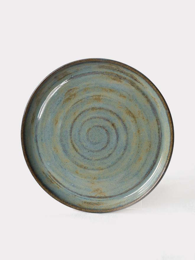 A handmade ceramic cake plate in seafoam blue glaze, photographed from the front against a plain light grey background, showing a natural concentric swirl texture and a shallow raised rim