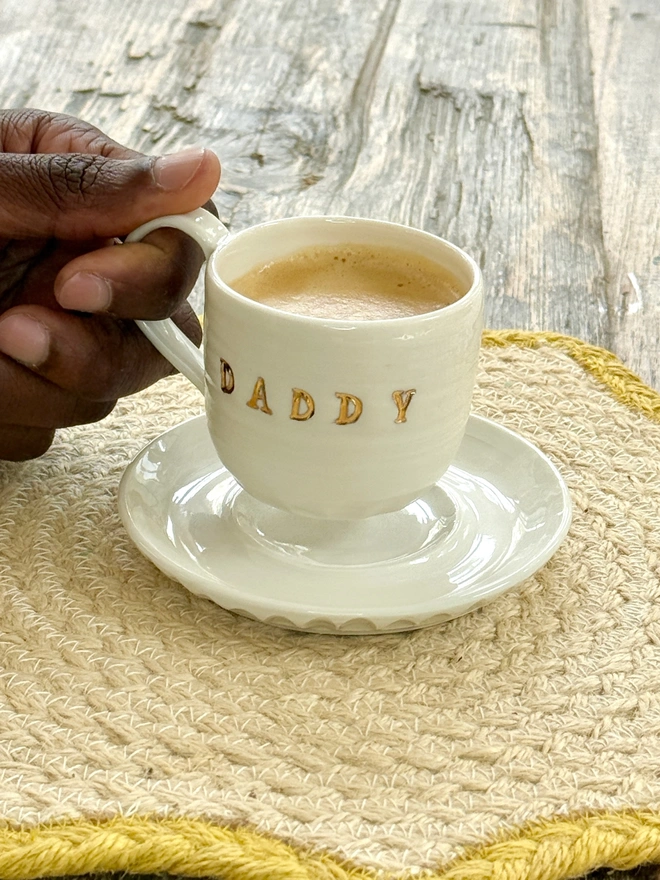 Espresso cup and saucer with DADDY emblazoned on the side with gold lustre detailing being lifted by a hand