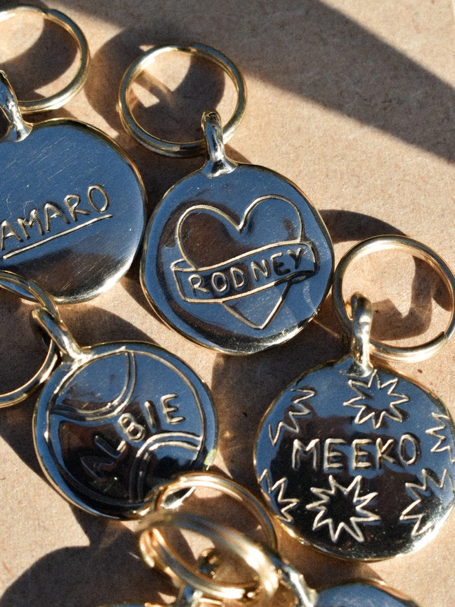 shiny brass round pet tags with an assortment of doodles, on says 'RODNEY' on a banner wrapped around a heart, one said 'ALBIE' inside a tennis ball and one says 'MEEKO' surrounded by stars, hand engraved. they lay in the sun on a pale brown background.