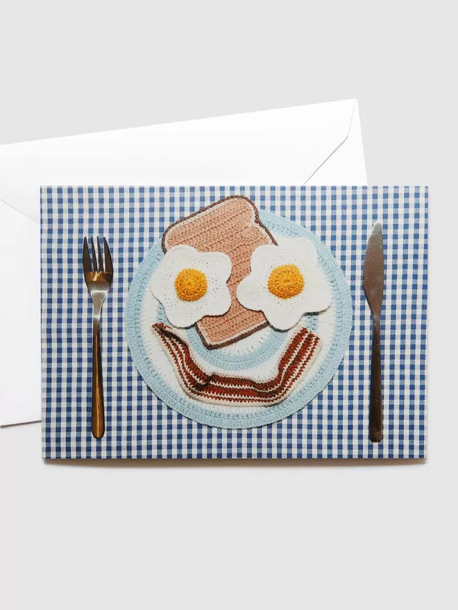Kate Jenkins Crispy Bacon Card seen with its white envelope. On the front are Kate's crotched eggs, bacon, toast and plate, organised as a smiley face. This is placed on a blue and white chequered tablecloth with a knife and fork either side.