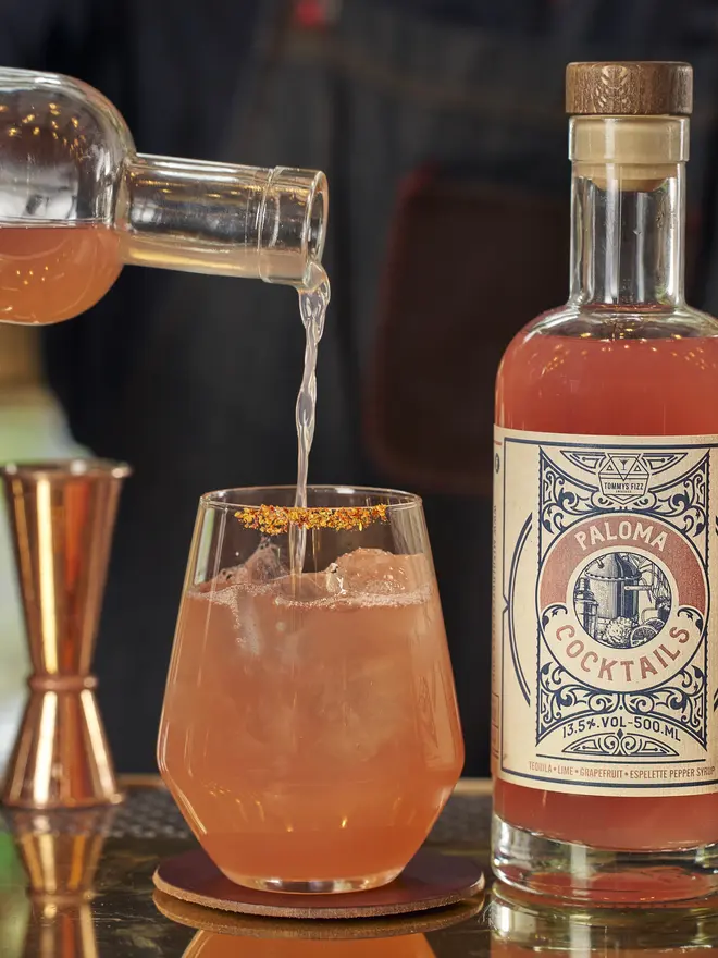 Tommy's Fizz Paloma Cocktail, a pink Paloma cocktail with a chili-salt rim and a lime wedge next to its bottle.
