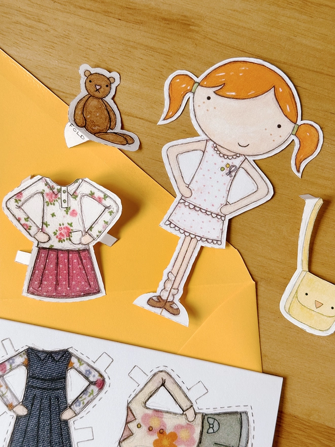 A paper doll greetings card with a paper doll and several outfits on the card lays on a yellow envelope on a wooden desk.