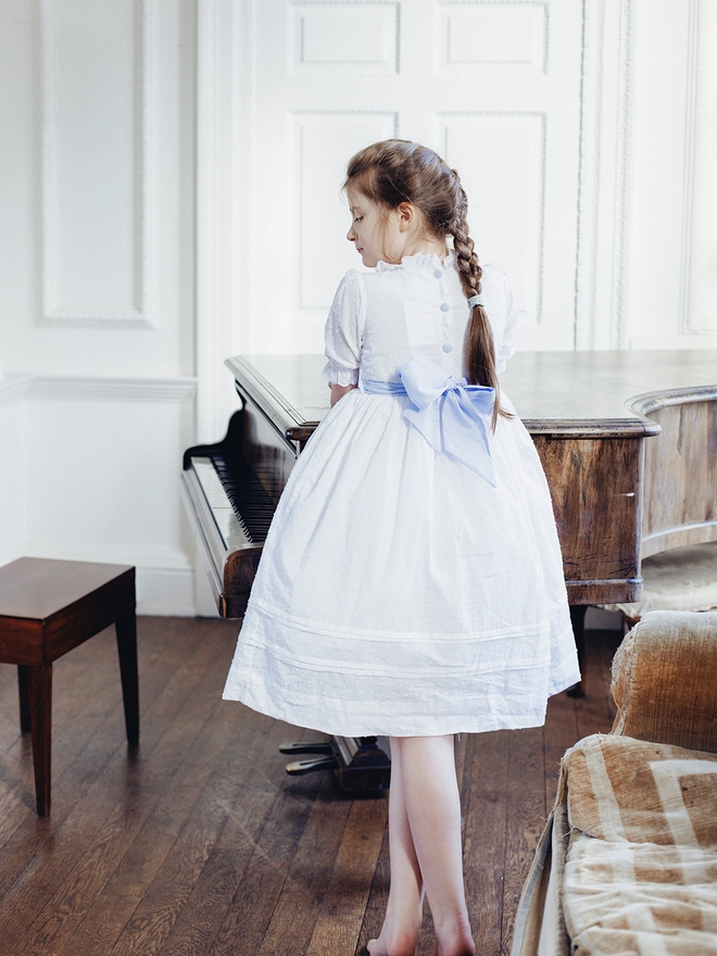 A girl stands leaning on a piano in a white dress with a blue sash