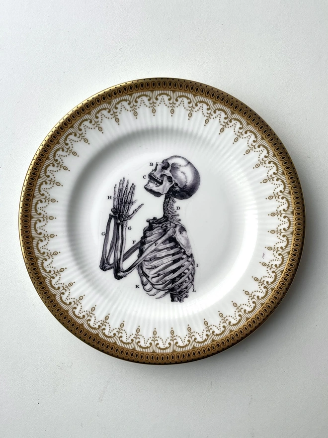 a vintage plate with an ornate gold border has a vintage illustraion in black and white of a praying skeleton in the centre of the plate