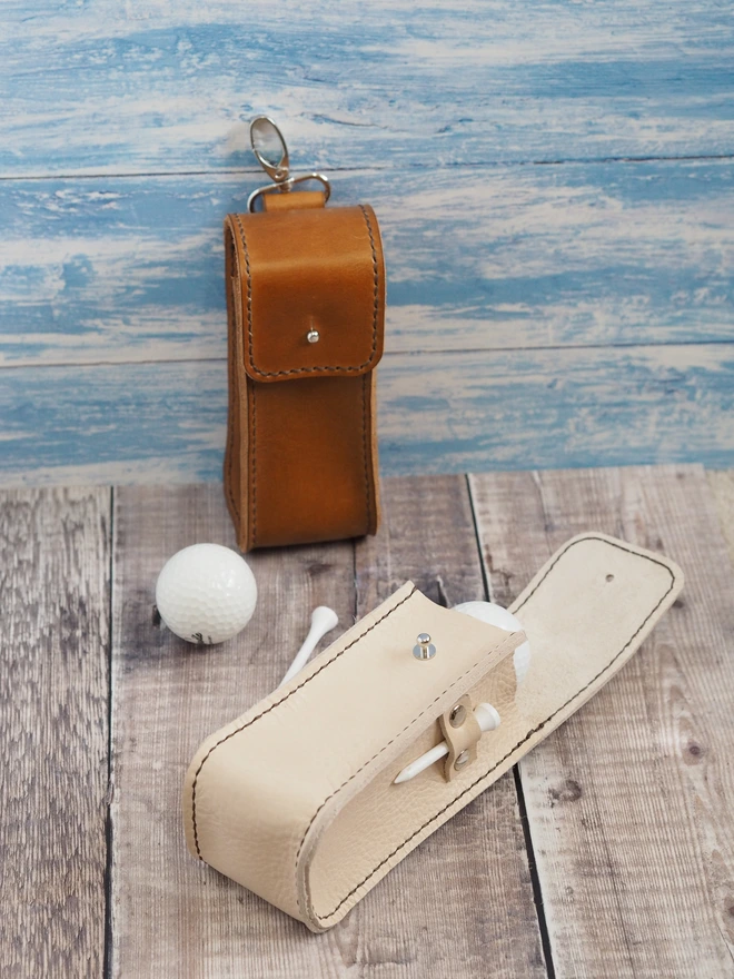 Handmade Leather Golf Ball Case. A hand dyed tan brown and natural cream leather pouch sit side by side, with golf ball and tee.