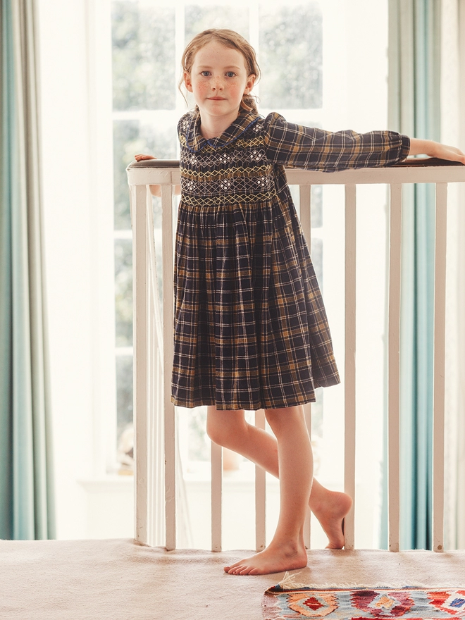 A girl hangs off railings wearing a navy and yellow dress with hand smocked detail