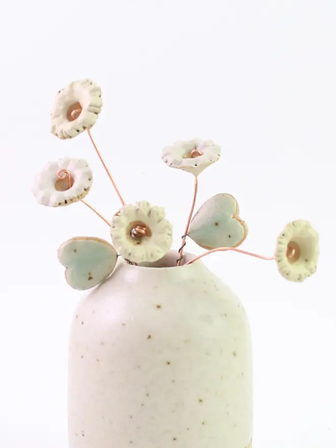 White and yellow pastel ceramic wildflowers with copper stems and two mint green heart leaves coming out of a speckled white ceramic vase