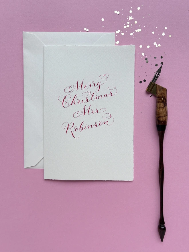 Christmas card and envelope on a pink background with a calligraphy pen.