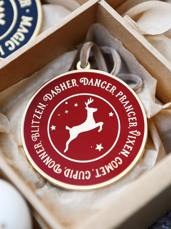A deep red and gold enamel Christmas decoration, with the names of all eight reindeer surrounding a gold reindeer, is tucked into a sectioned box.