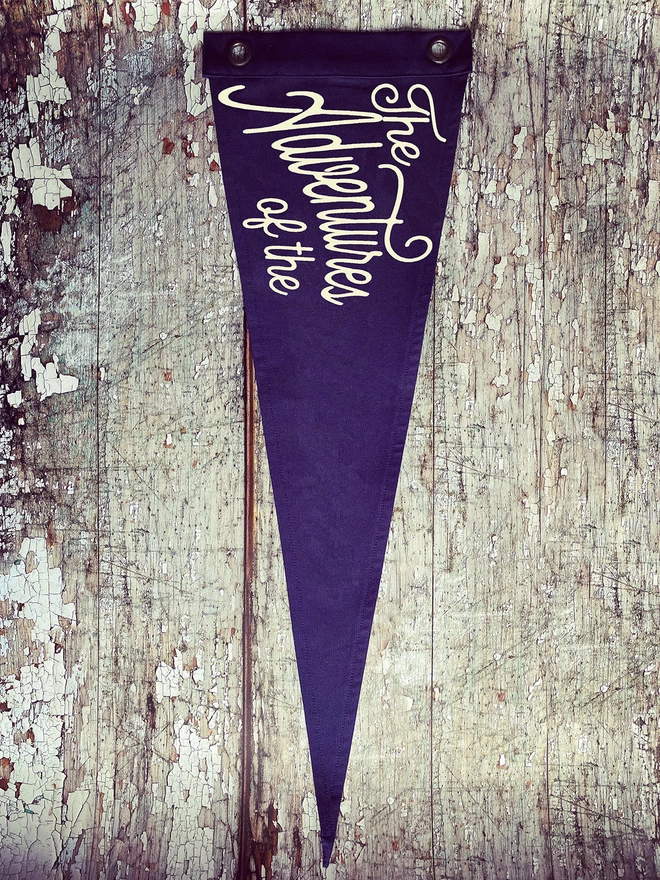 A navy coloured pennant flag hung vertically on a distressed wooden wall. In ivory lettering is the words ‘The Adventures of the’