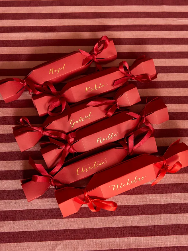 A set of 6 personalised Christmas crackers in red with gold calligraphy with matching ribbon, atop a red and light pink striped tablecloth