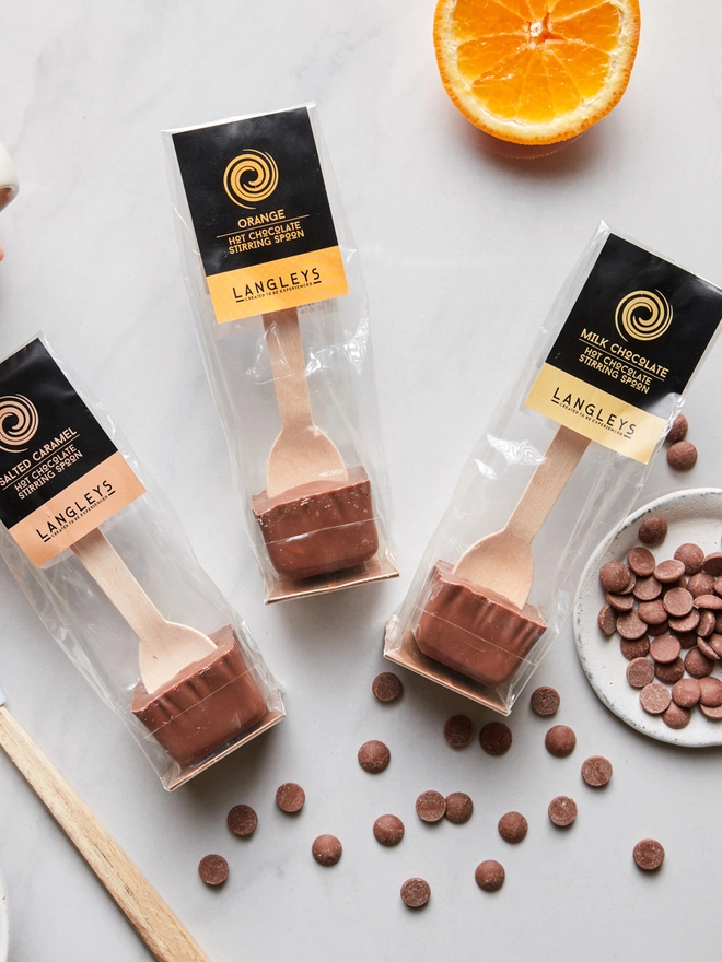 Hot Chocolate Stirring Spoon Collection (Set Of 3), a group of chocolate serving spoons sitting on a plain surface. They are accompanied by other edible items such as fruit and smaller chocolates. 