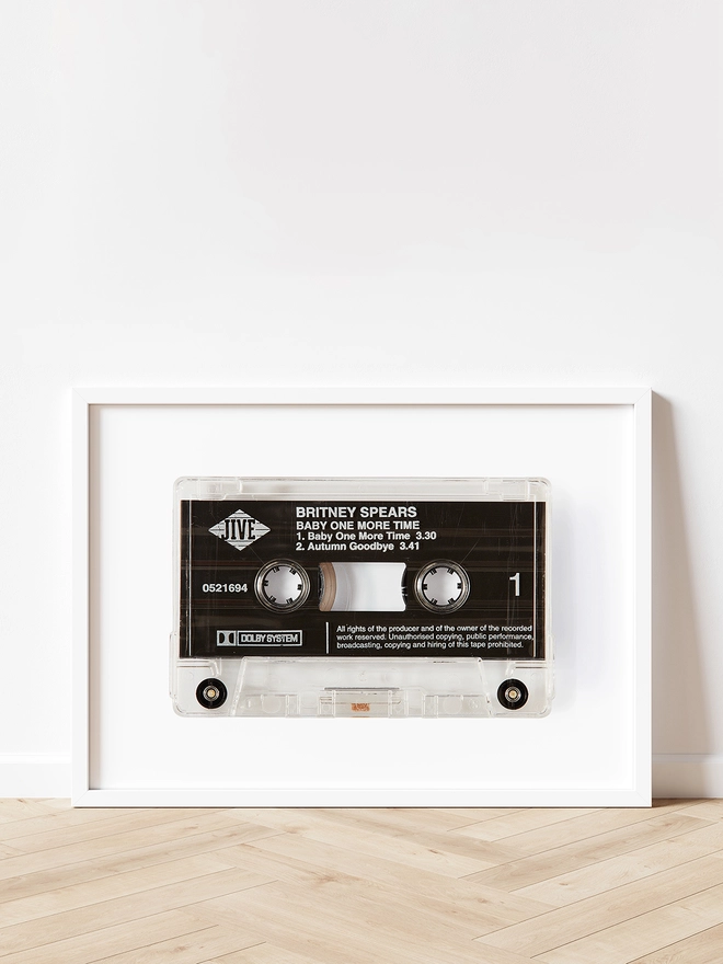 baby one more time britney spears cassette print. framed print propped up against white wall on wooden floor.