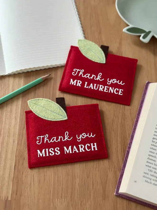 Two handmade red felt apple-shaped envelopes with green felt leaves and brown stems, each reading "Thank you MR LAURENCE" and "Thank you MISS MARCH", displayed on a wooden desk with a pencil, notebook, and book. It's such a sentimental personalised, handmade teacher thank you gift.