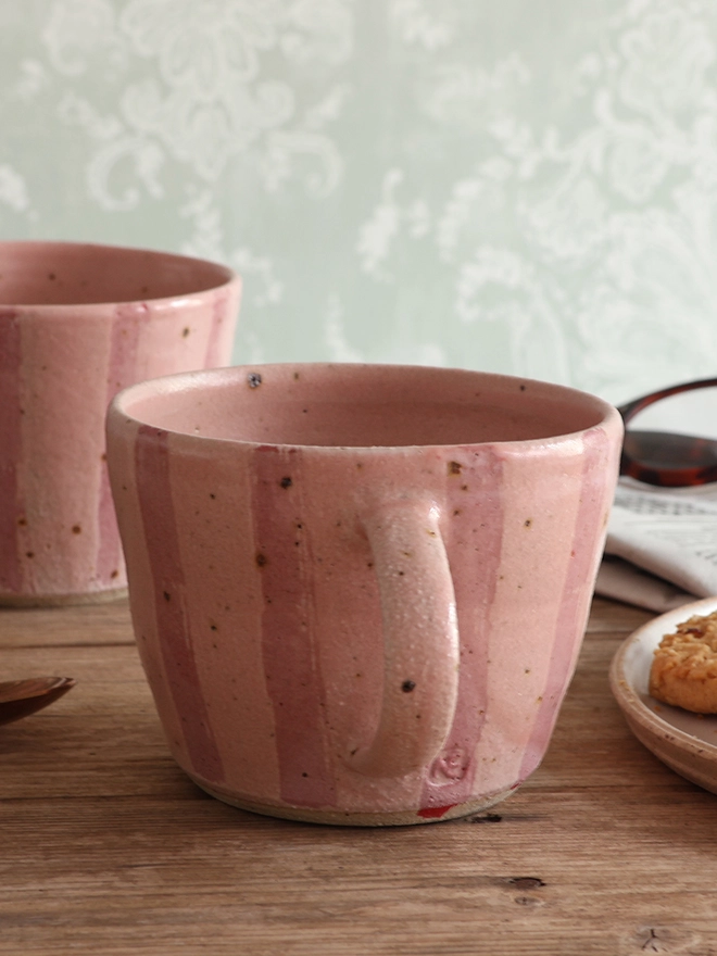 Handmade ceramic stoneware mug in pink stripe sat on a wooden surface with another mug in the same style sat behind it. The background also shows a newspaper with glasses on top, a brass spoon and a plate with cookies. 