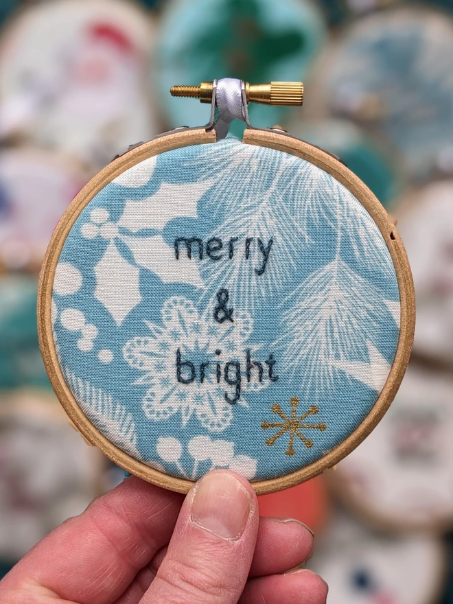 A hand embroidered hoop on Christmas fabric reading merry & bright