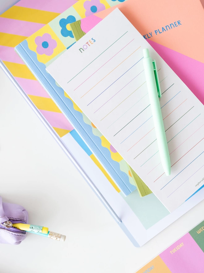 The Raspberry Blossom rainbow coloured lined note pad sits on a desk in a pile of other colourful stationery items from the Happiness collection