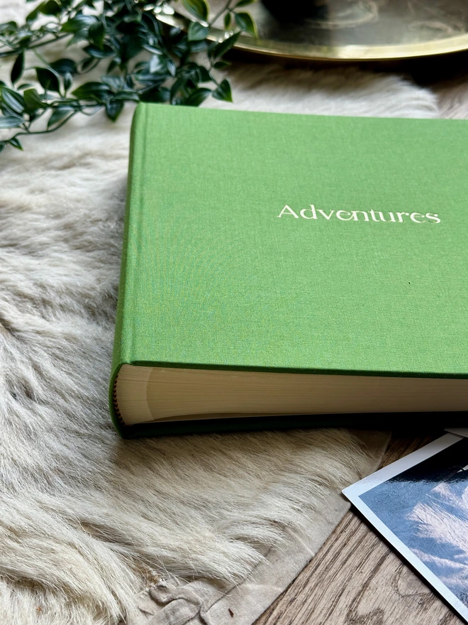 A hand-bound green photo album printed with the words 'Adventures' in gold lettering rests on a bookshelf next to rows of red books.