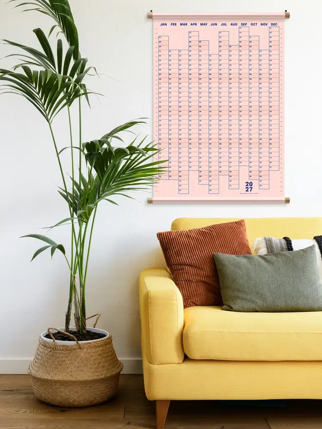 A pink 'nude' Dotline 2027 yearly wall planner on a white wall, with plant to the side and yellow sofa with cushions underneath