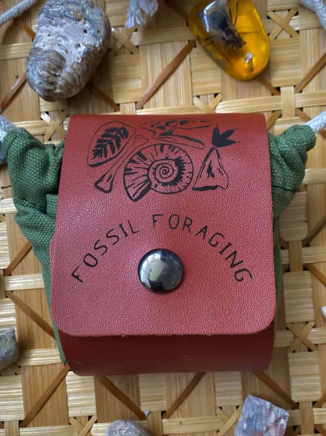 Personalised Foraging Drawstring Canvas Pouch, a green canvas pouch with a brown leather flap that reads "FOSSIL FORAGING" surrounded by rocks, fossils, and amber on a woven mat.