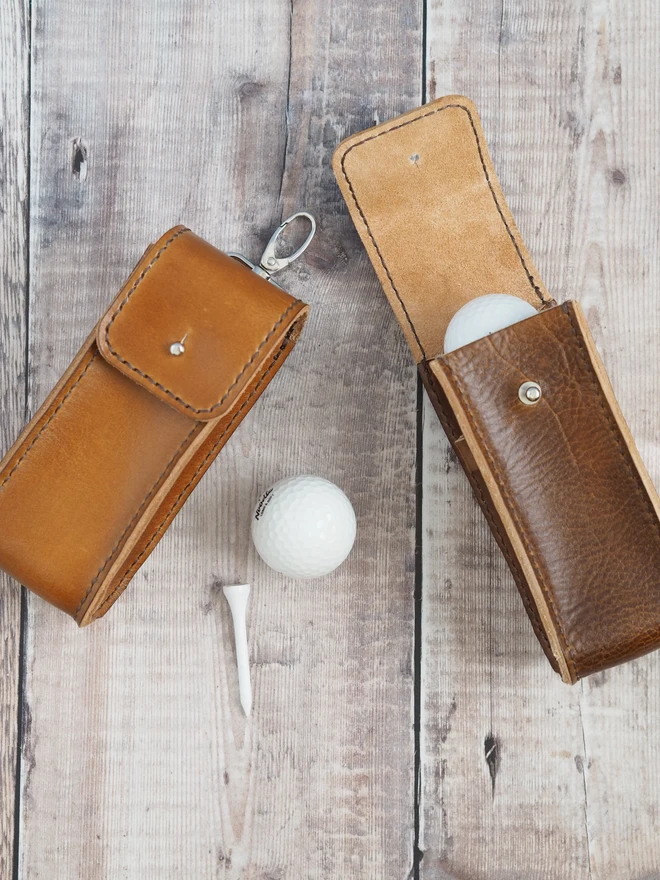 Hand dyed tan brown leather golf ball carry pouch. Two brown cases sit together, one is a darker brown and the other slightly lighter, the natural patina of handmade goods.