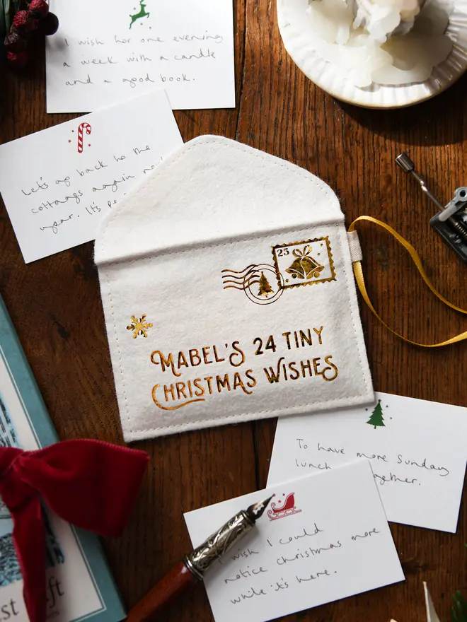 A handmade felt envelope advent calendar with golden lettering that reads 'Mabel's 24 Tiny Christmas Wishes' with several Christmas notecards all laying on a wooden table.