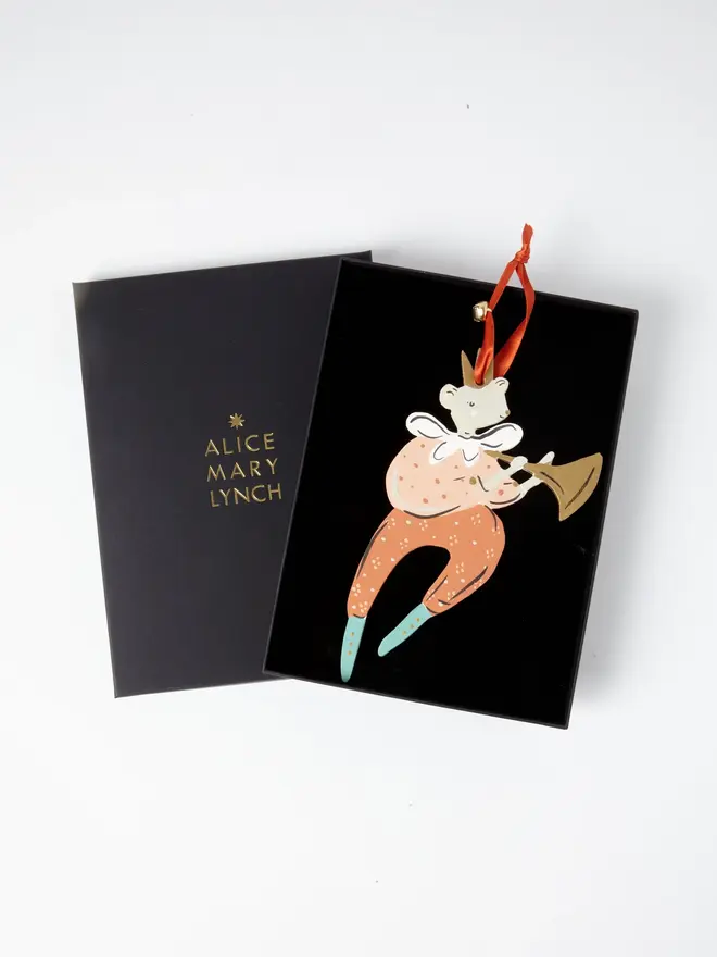 Mouse Herald Tree Dancer. the mouse herald tree dancer hanging ornament in coral and peach tones sits inside its black presentation box, with the lid open to reveal "alice mary lynch" embossed in gold lettering. the mouse figure wears a gold crown and a ruffled white collar, playing a gold trumpet, with spotted coral trousers ending in teal boots. the branded box gives the piece a polished, gift-ready presentation.