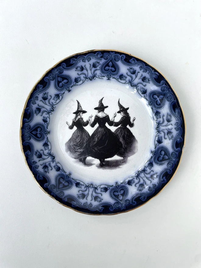 a coven of 3 witches dance hand in hand, the image is victorian in black and white and is printed directly onto an ornate vintage plate