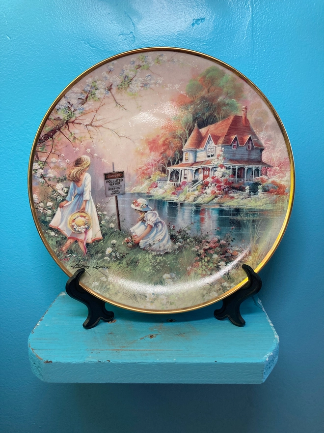 Beautiful decorative 8” plate featuring a watercolour style image of two young girls playing by a river. On the other side of the river is a beautiful white house. The scene is idyllic, yet in the river we see a warning sign which says ‘Danger! Polluted Water’...