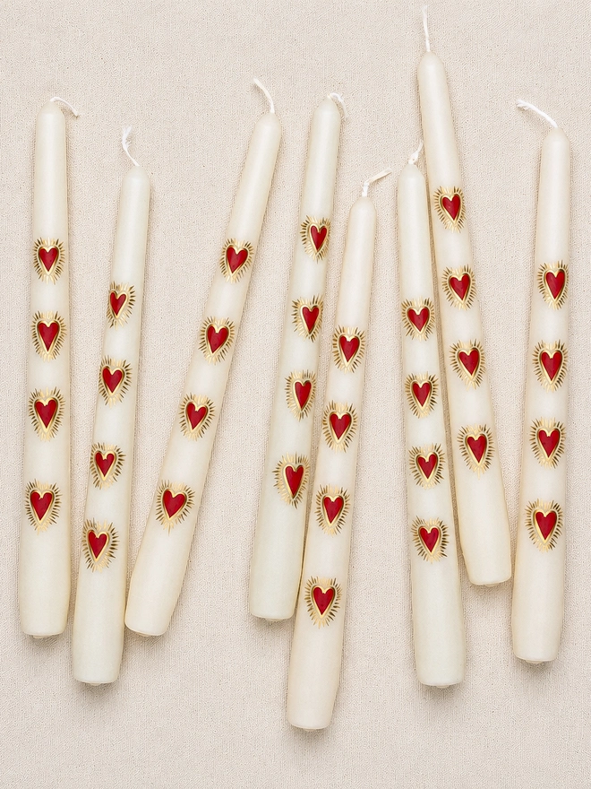 Eight ivory hand-painted taper candles with red sacred heart motifs and gold detailing scattered across a soft cream linen background.