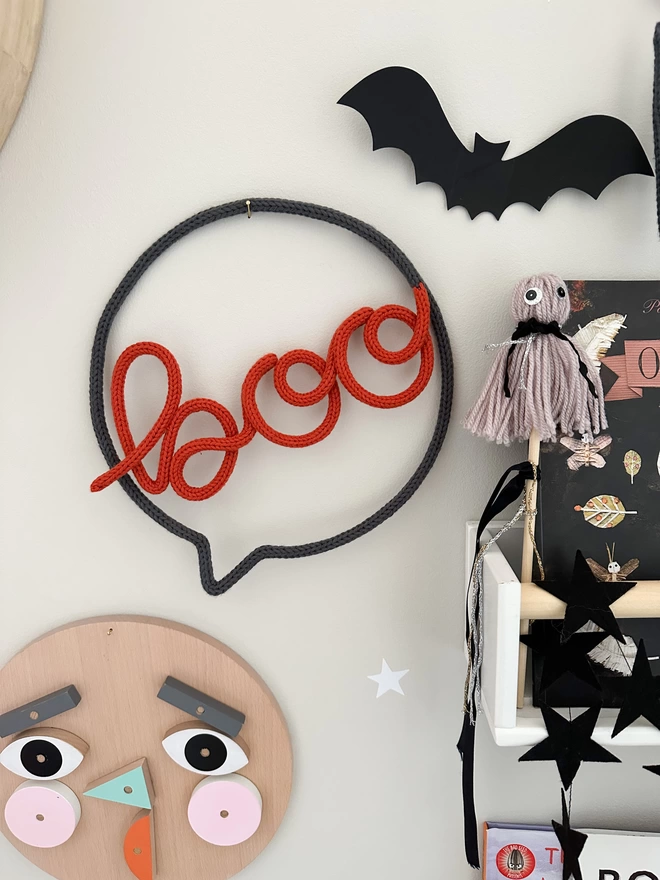 'boo' speech bubble Kid's Halloween Knitted Wall Decoration in colours 'fox' and 'graphite' hanging up on the wall above a wooden face wall hanging.